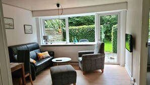 A living room with a sofa, armchair, ottoman, and a small table. There is a window with a view of greenery outside. - Sea sun (Nørre Nebel)