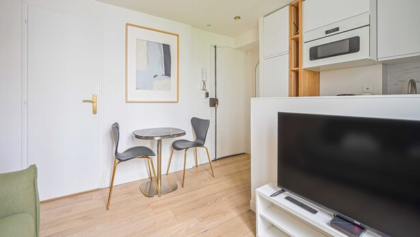 A modern living room with a flat-screen TV, a dining table with two chairs, and a kitchen area with white cabinets and a microwave. - Cosy Nest - 2P - The 16th Arrondissement (Paris)