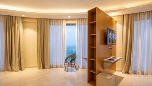 Una habitación de hotel moderna con techo curvo, un televisor montado en un estante de madera, una silla y vistas de la ciudad a través de amplios ventanales.