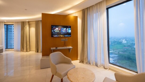 Una habitación de hotel moderna con un televisor grande montado en la pared, una zona de estar cómoda con una mesa redonda y un ventanal con vistas a la ciudad.