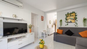 Un salón con un televisor de pantalla plana, un sofá gris con cojines anaranjados, un mueble de televisión blanco y una mesita con un jarrón amarillo.