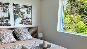 Een slaapkamer met een bed, kussens, een kunstwerk aan de muur en een raam met uitzicht op groen.