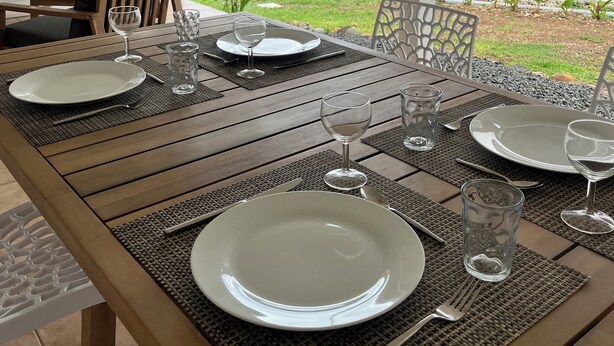 Een houten eettafel gedekt met witte borden, bestek en wijnglazen, onder een witte pergola met uitzicht op een weelderige tuin en een huis op de achtergrond.