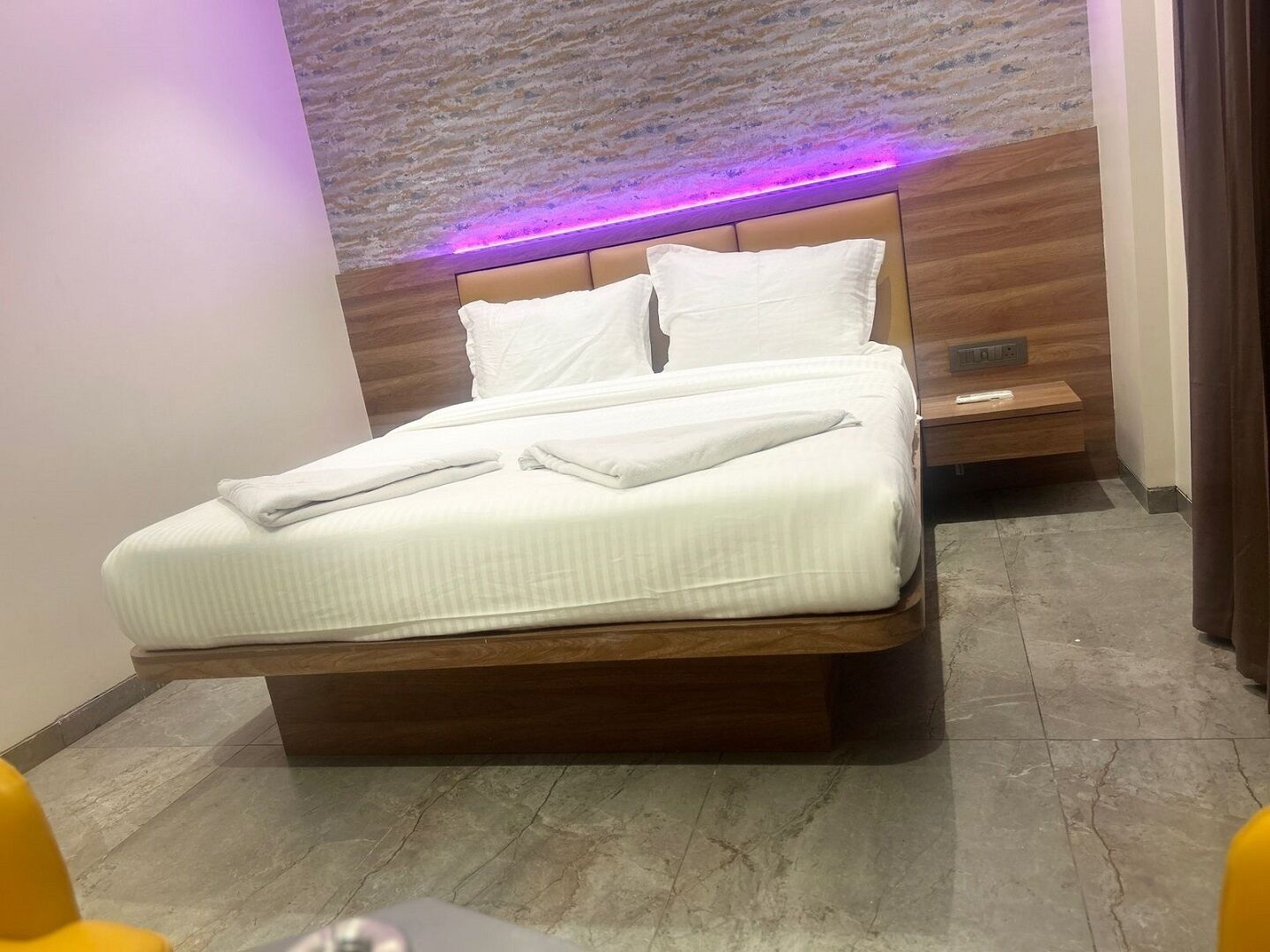 Een net opgemaakt bed met witte lakens en handdoeken, een houten hoofdbord en een nachtkastje met een stopcontact.