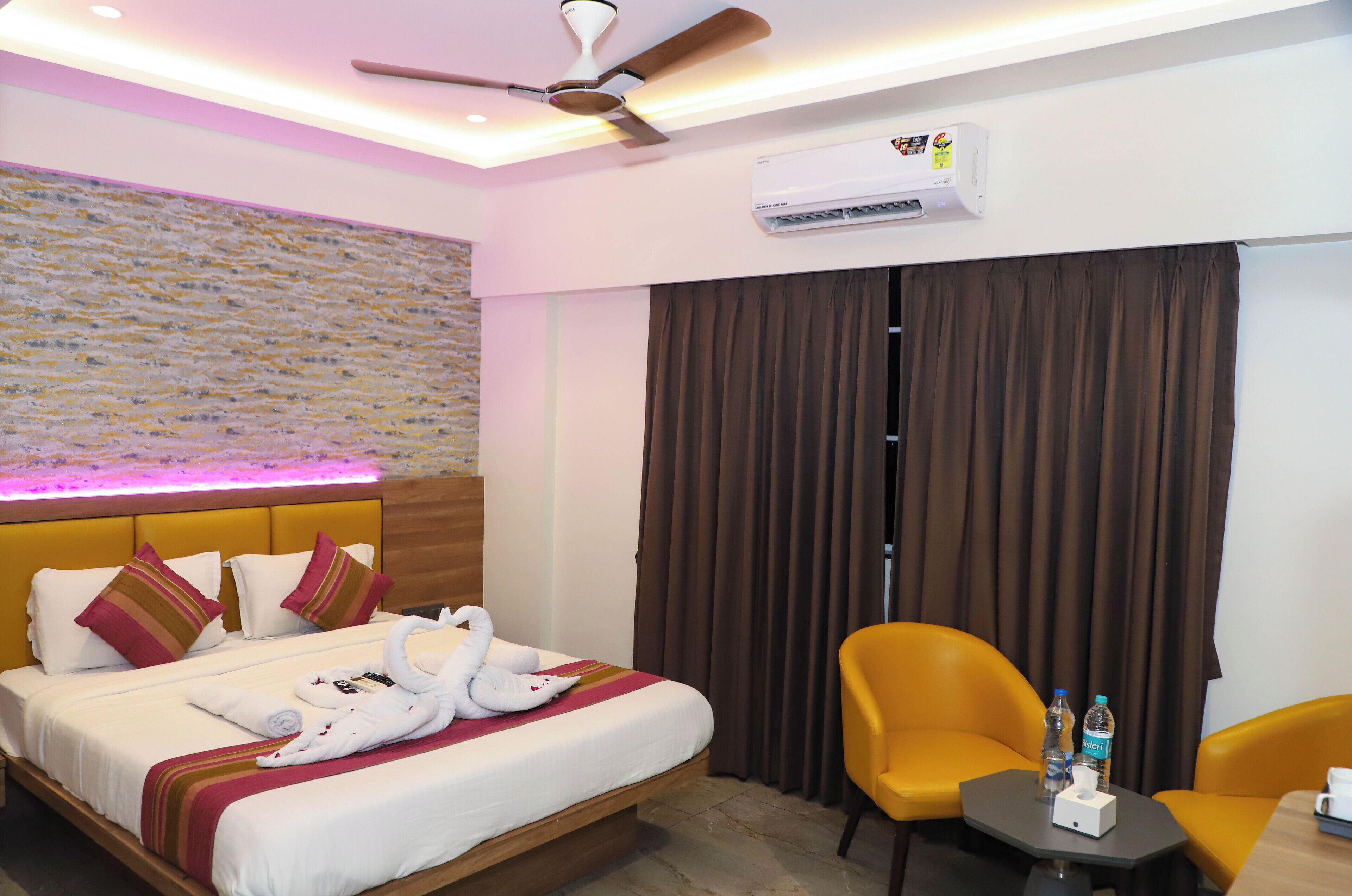 A hotel room with a bed, a ceiling fan, an air conditioning unit, a brown curtain, a small table with a bottle and a cup, and a yellow chair.