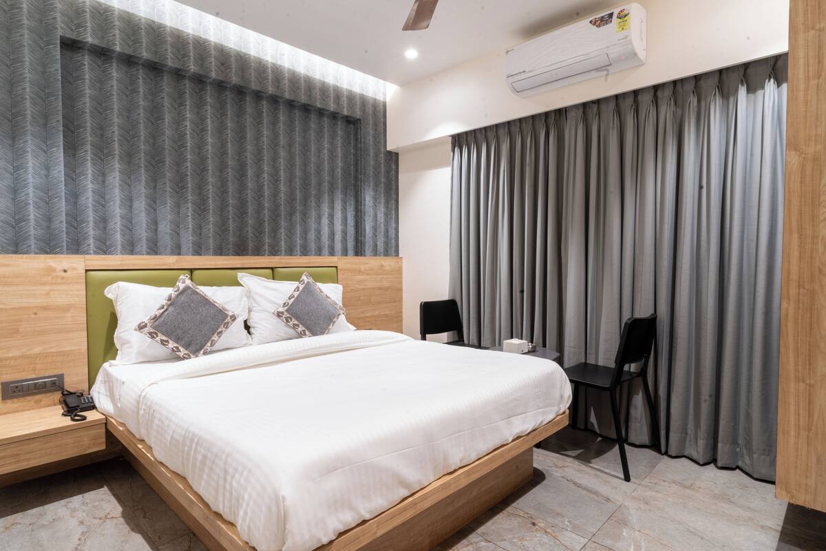 Quarto de hotel moderno com cabeceira de madeira, uma cama grande com roupa de cama branca, uma escrivaninha com cadeira e uma janela com cortinas cinzas.