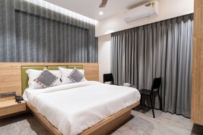 A modern hotel room with a wooden headboard, a large bed with white linens, a desk with a chair, and a window with grey curtains. - Hotel Aura Inn (Navi Mumbai)