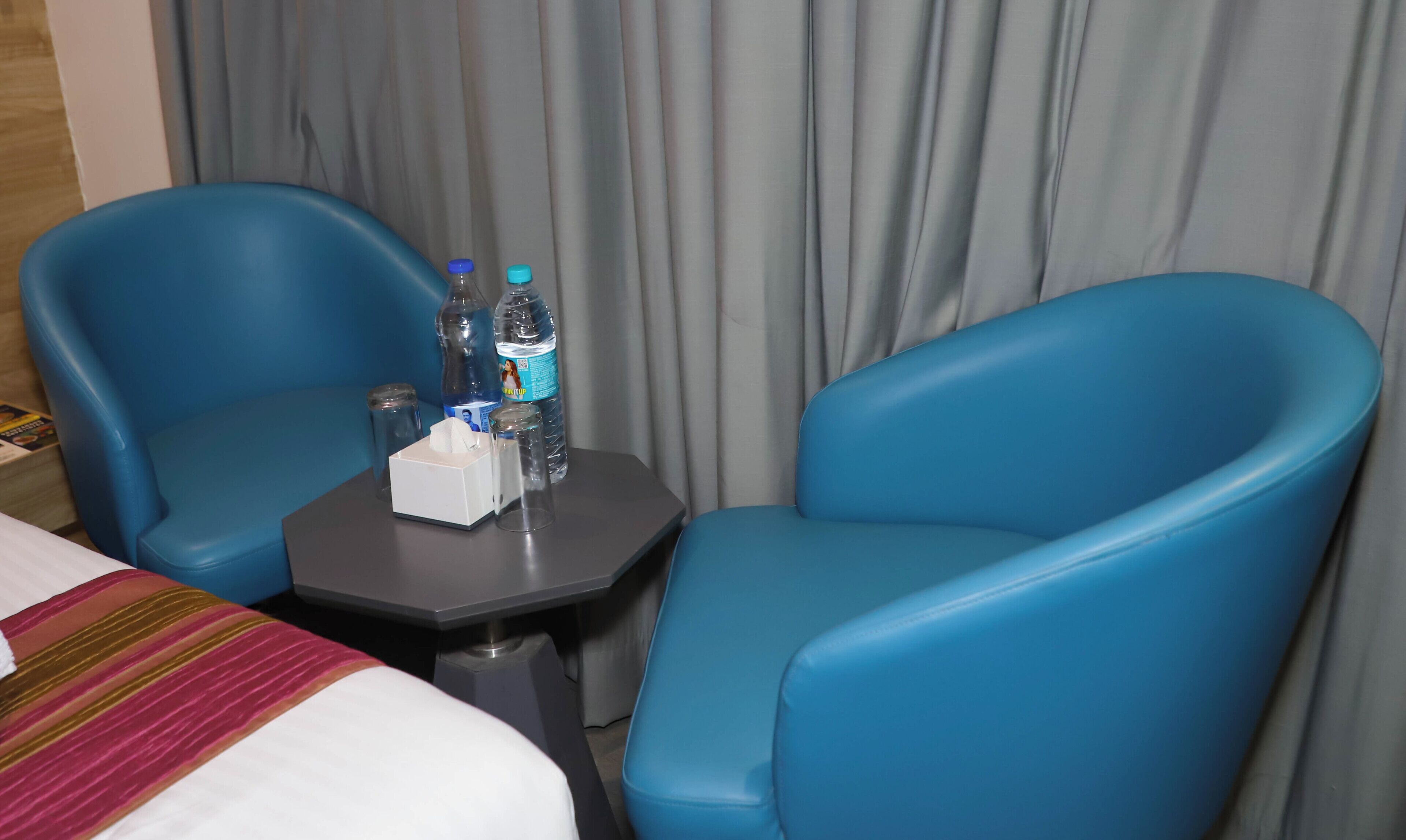 A hotel room with two blue armchairs, a small table with water bottles and a tissue box, and a bed with a striped blanket.