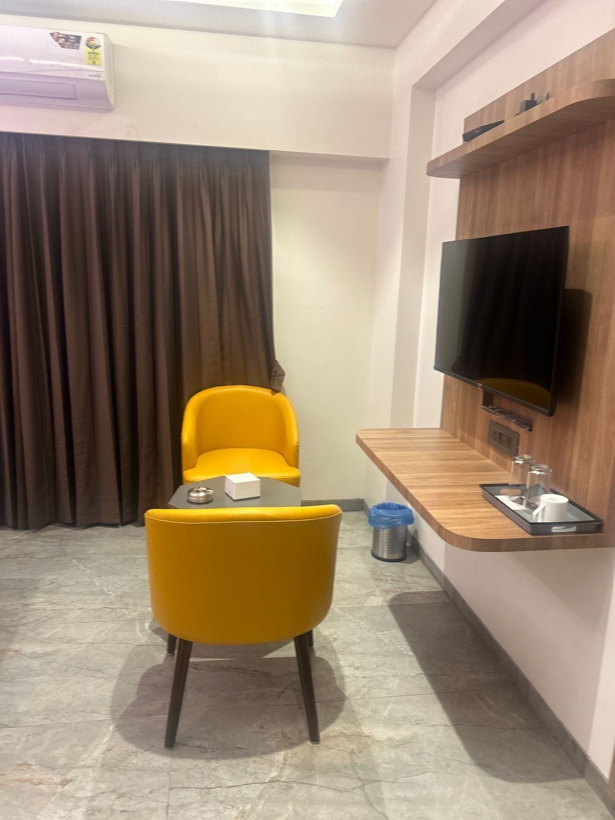 A hotel room with a dining area featuring a table and two yellow chairs, a TV mounted on a wooden wall, and a window with curtains.