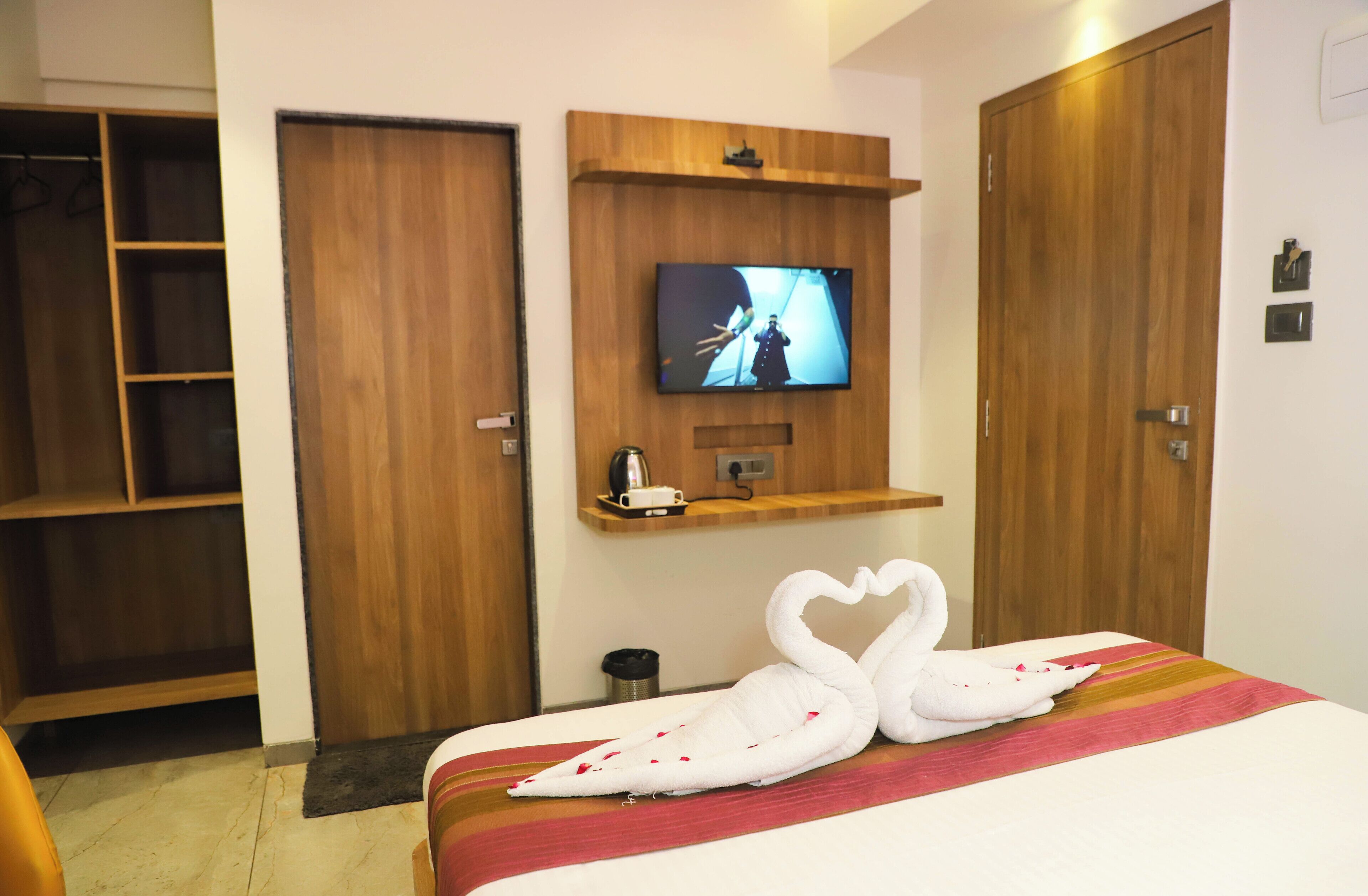 A hotel room with a bed, a TV mounted on the wall, a wooden wardrobe, and a shelf with a kettle and a phone.