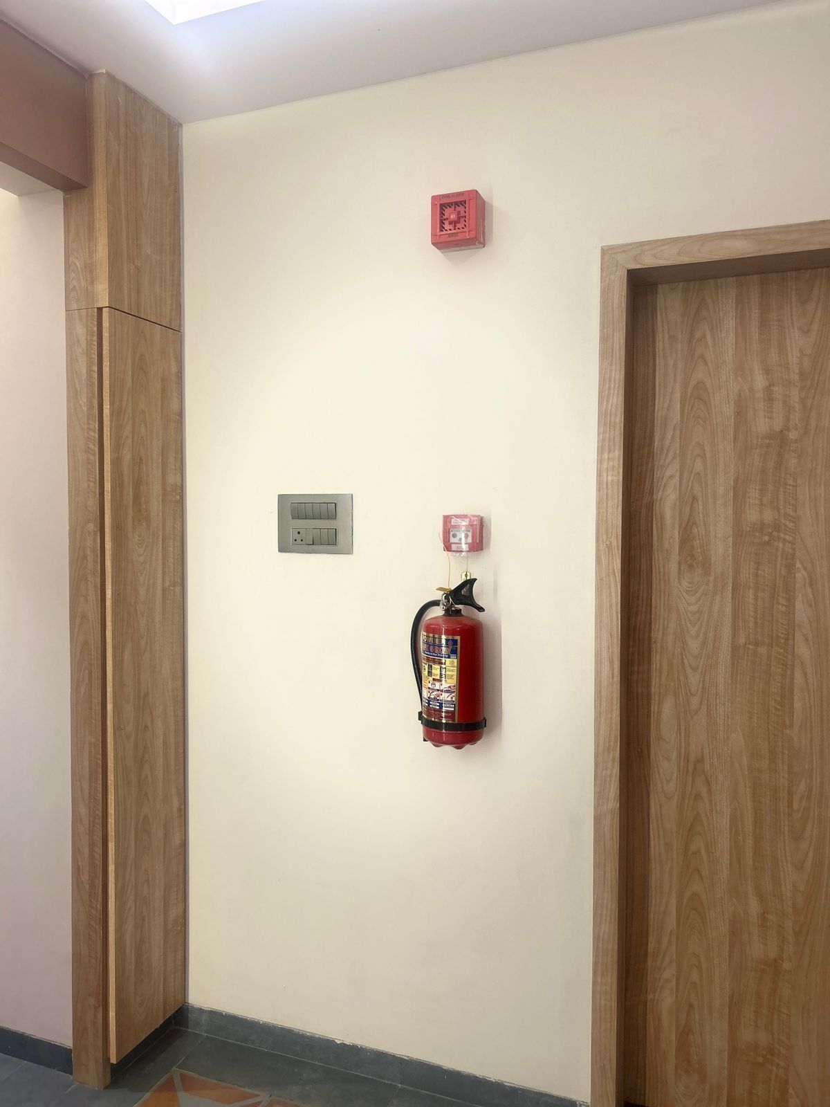 Un pasillo con un extintor montado en la pared, un enchufe y una pequeña caja roja.
