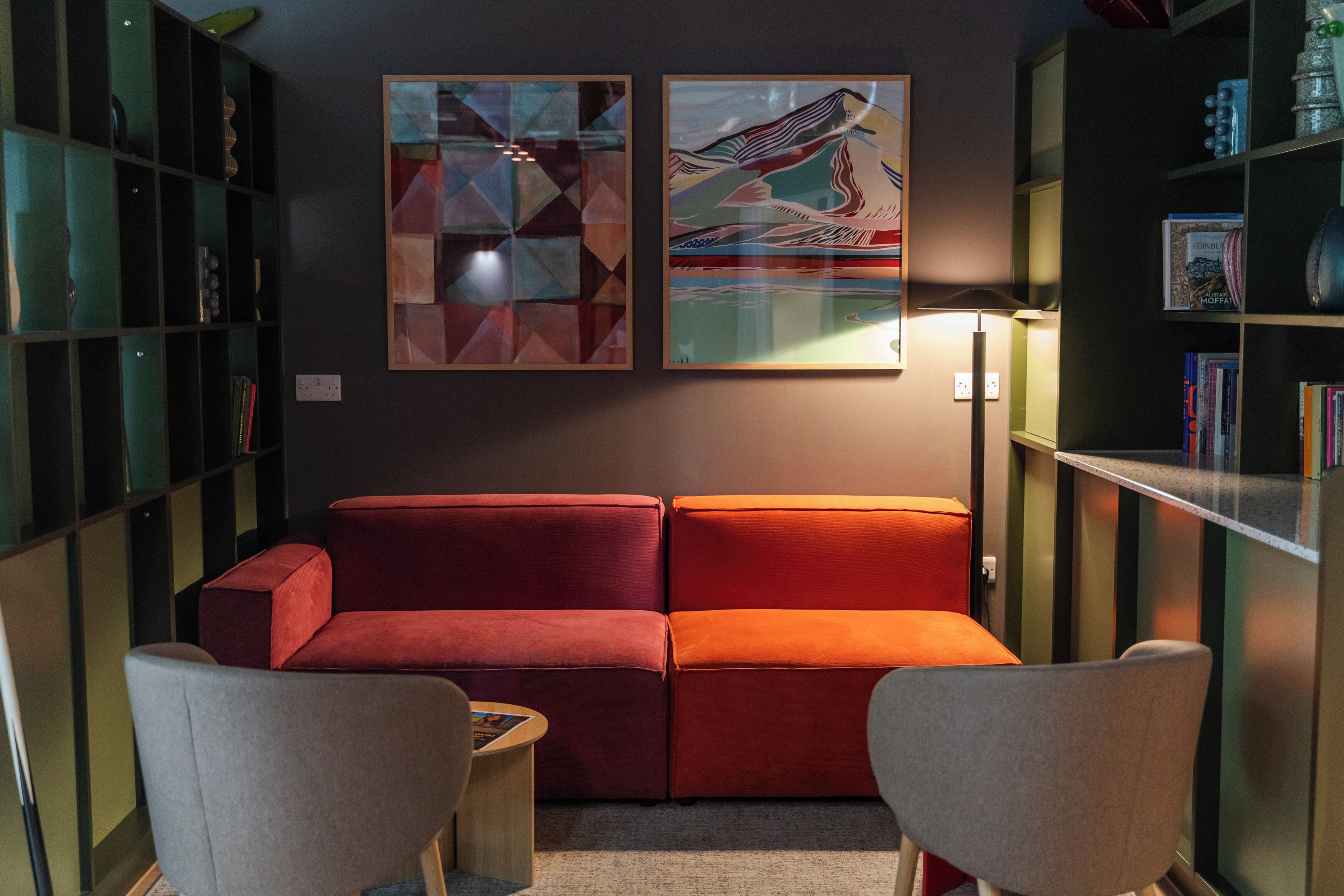 Un salon moderne avec un canapé rouge, deux fauteuils et une table basse. Les murs sont ornés de tableaux abstraits, et une bibliothèque remplie de livres et d’objets décoratifs se trouve dans la pièce.