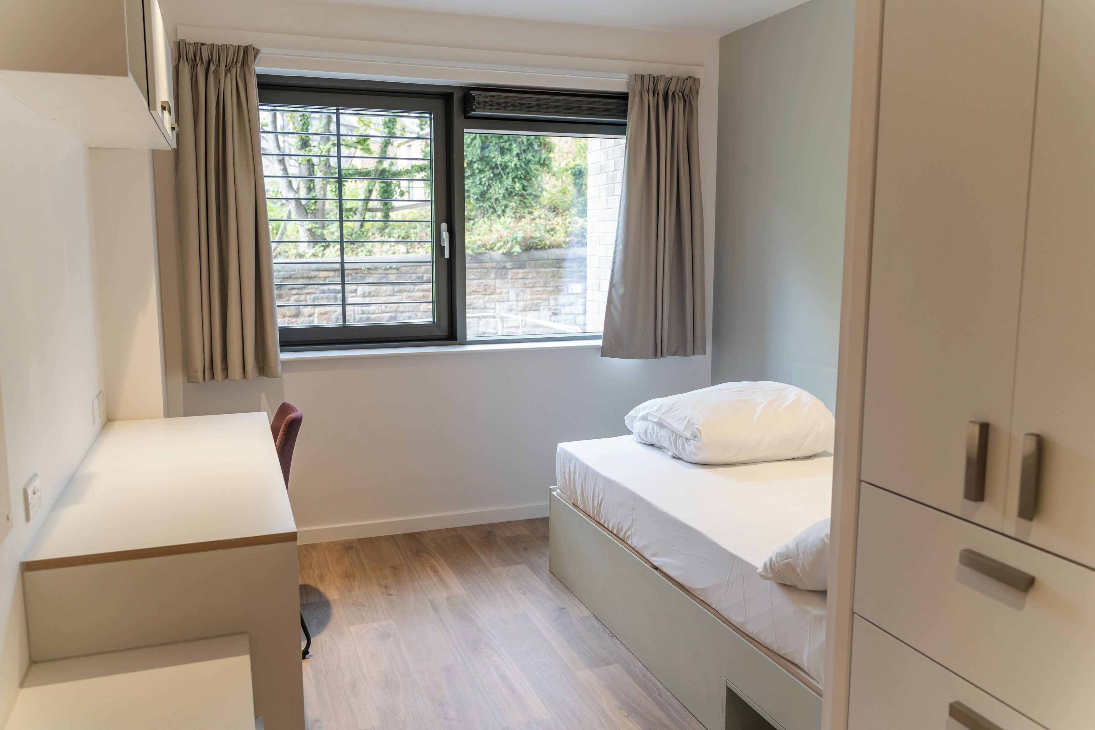 A small, modern bedroom with a single bed, a desk, and a window with curtains.