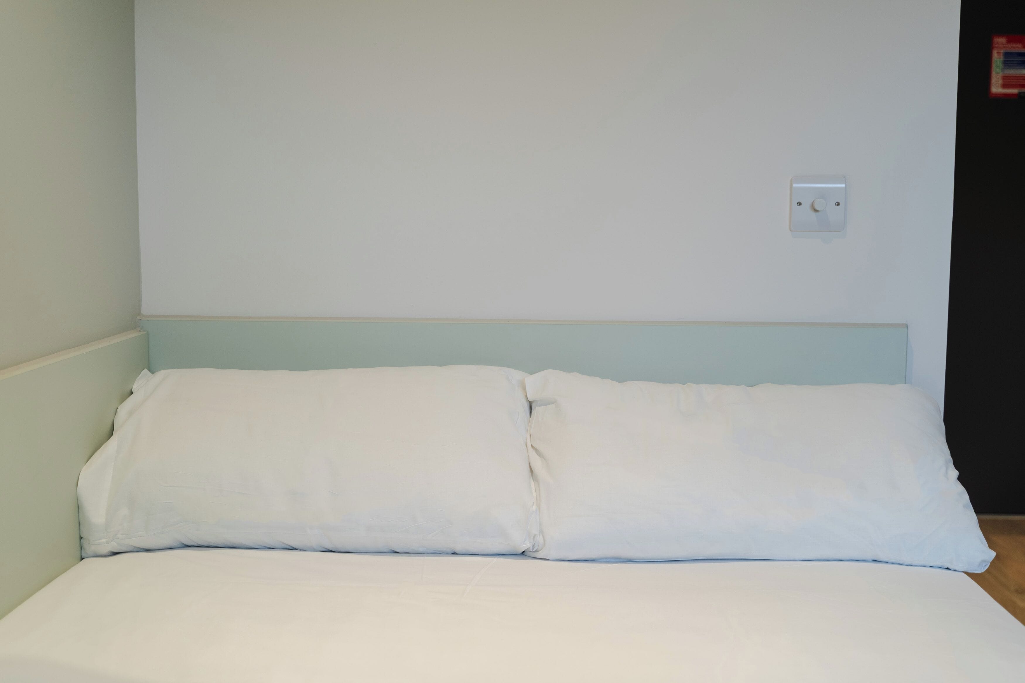 A simple, unadorned bed with white bedding in a room with light blue walls and a plain white ceiling.