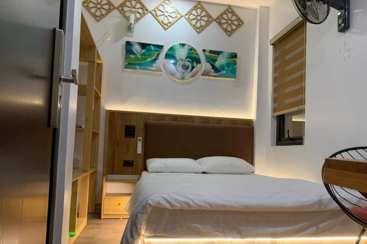A bedroom with a bed, wooden headboard, bedside tables, a wall-mounted artwork, and a fan.