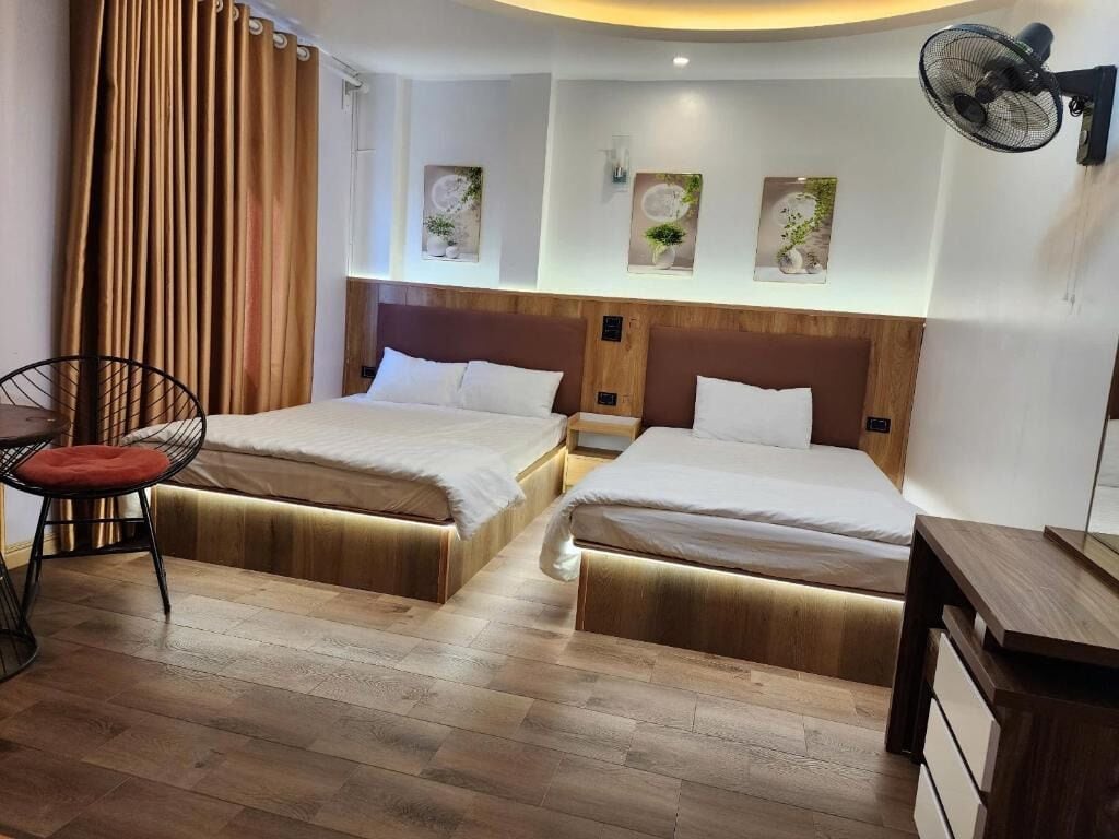 A hotel room with two beds, a wooden floor, a desk with a chair, and a fan.