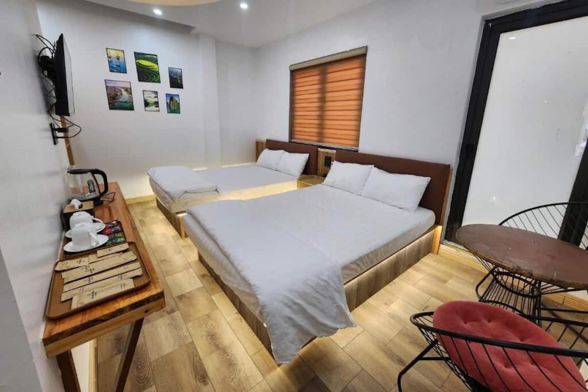 A hotel room with two beds, a wooden table with a tray of teapots, a red chair, and a window with blinds.