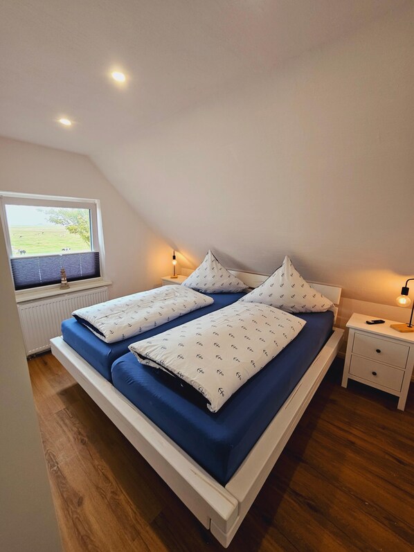 A bedroom with a sloped ceiling, a window with blinds, a bed with blue bedding, two bedside tables with lamps, and a wooden floor.