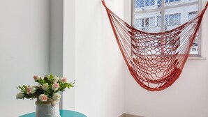 Una habitación de esquina con una hamaca, una mesita con un florero, un cuadro enmarcado y una ventana con vistas a edificios.