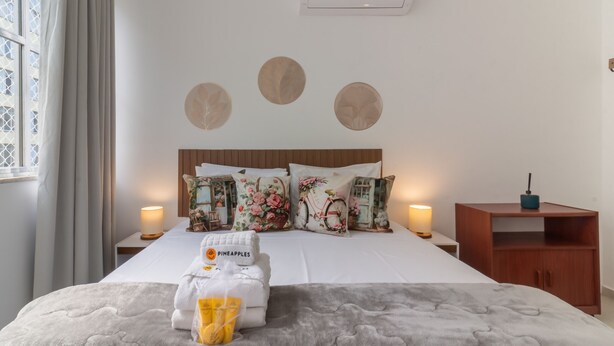 Una habitación de hotel con una cama bien hecha, almohadas estampadas con motivos florales, un cabecero de madera, una mesita de noche con lámpara y un pequeño mueble de madera.
