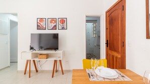 Un comedor con una mesa de madera dispuesta para dos personas, un televisor sobre un soporte blanco, y una puerta de madera.