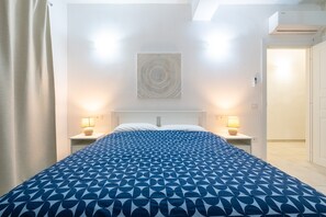 Ett hotellrum med en säng, två sänglampor, ett konstverk på väggen och en dörr som leder till ett annat rum.