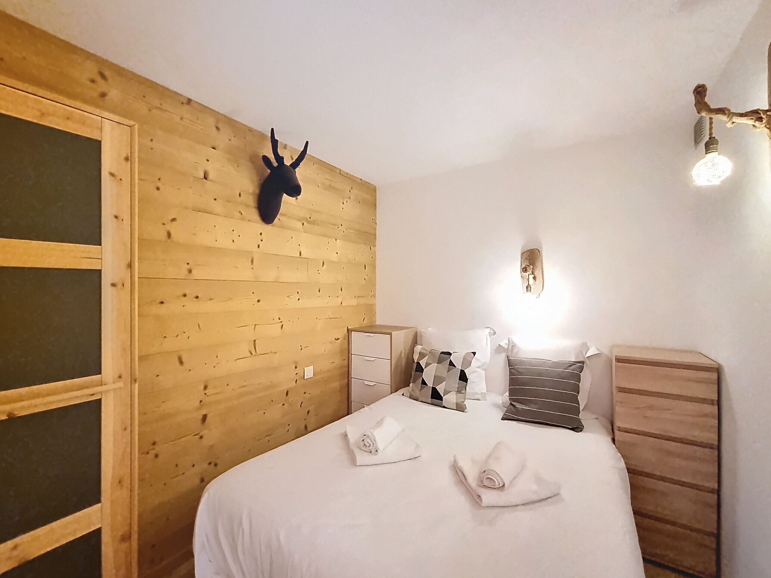 Et hotelværelse med trævæg, dekoration i form af et gevir, en seng med hvide sengetøj, et natbord, en kommode og en vægmonteret lampe.