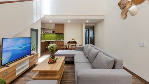 A modern living room with a flat-screen TV, a grey sectional sofa, a wooden coffee table, and a kitchen in the background. - Fusia Hotel & Apartment (Da Nang)