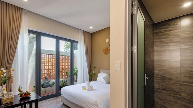 A hotel room with a bed, a desk with a vase, a window with curtains, and a view of the outside. - Fusia Hotel & Apartment (Da Nang)