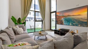 A modern living room with a large window, a sofa, a coffee table, and a painting of a coastal scene. - Spacious 2 BR + huge private garden in JVC (Jumeirah Village)