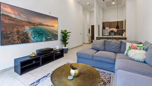 A modern living room with a large flat-screen TV, a grey sofa, a coffee table, and a potted plant. - Spacious 2 BR + huge private garden in JVC (Jumeirah Village)