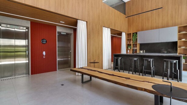 A modern interior with a long wooden table, bar stools, a kitchen area with a counter and stools, and two elevators. - 360 Pamplona NR  (São Paulo)