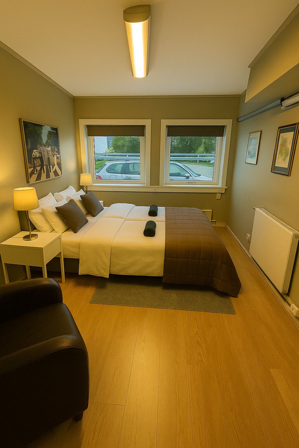 A bedroom with a bed, bedside tables, a lamp, a radiator, and windows with a view of cars outside.