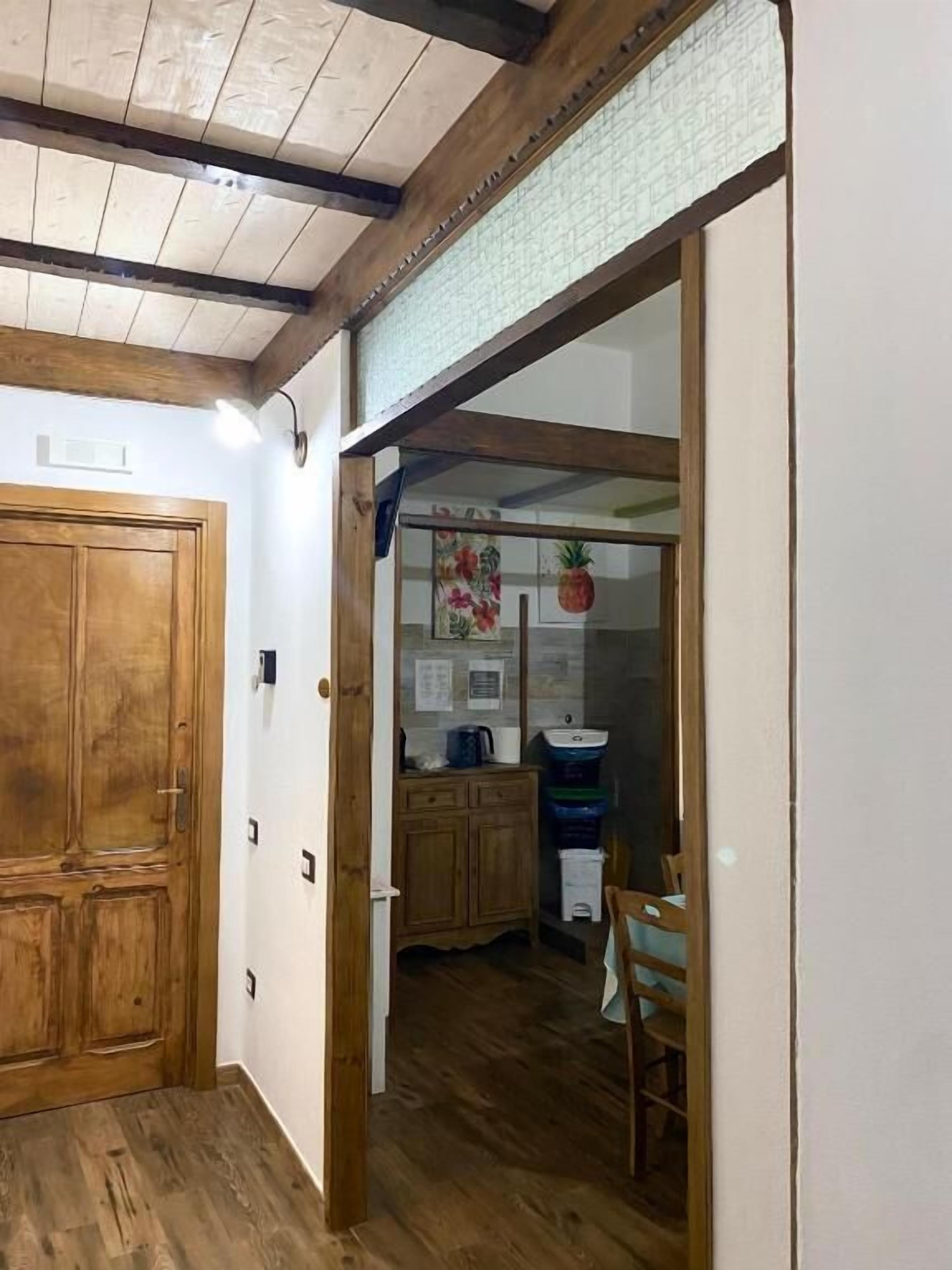 A hallway with wooden flooring, a wooden door, and a view into a kitchen area with a wooden table, chairs, and a hanging pineapple decoration.