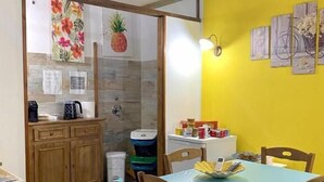 A dining area with a wooden table and chairs, a yellow wall, and a kitchen area with wooden cabinets and a fruit-themed painting. - Dani's House - affittacamere (Cagliari)