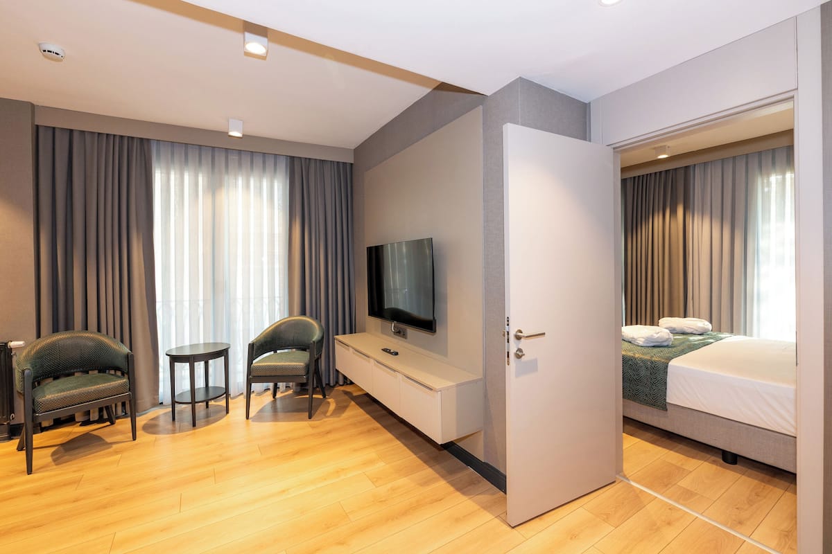 Quarto de hotel com cama, televisão, duas cadeiras, uma mesinha e cortinas.