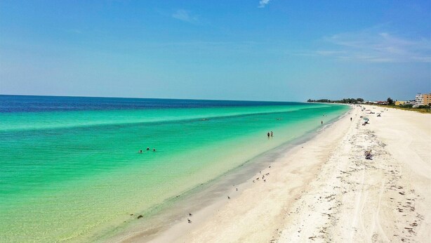Een breed zandstrand met helder turquoise water, enkele mensen die zwemmen en gebouwen op de achtergrond.