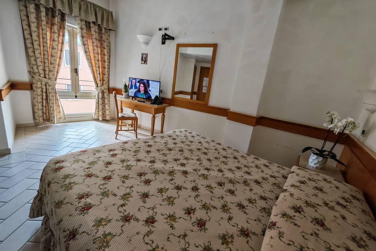 A hotel room with a bed, a desk with a computer, a chair, a mirror, and a window with curtains.