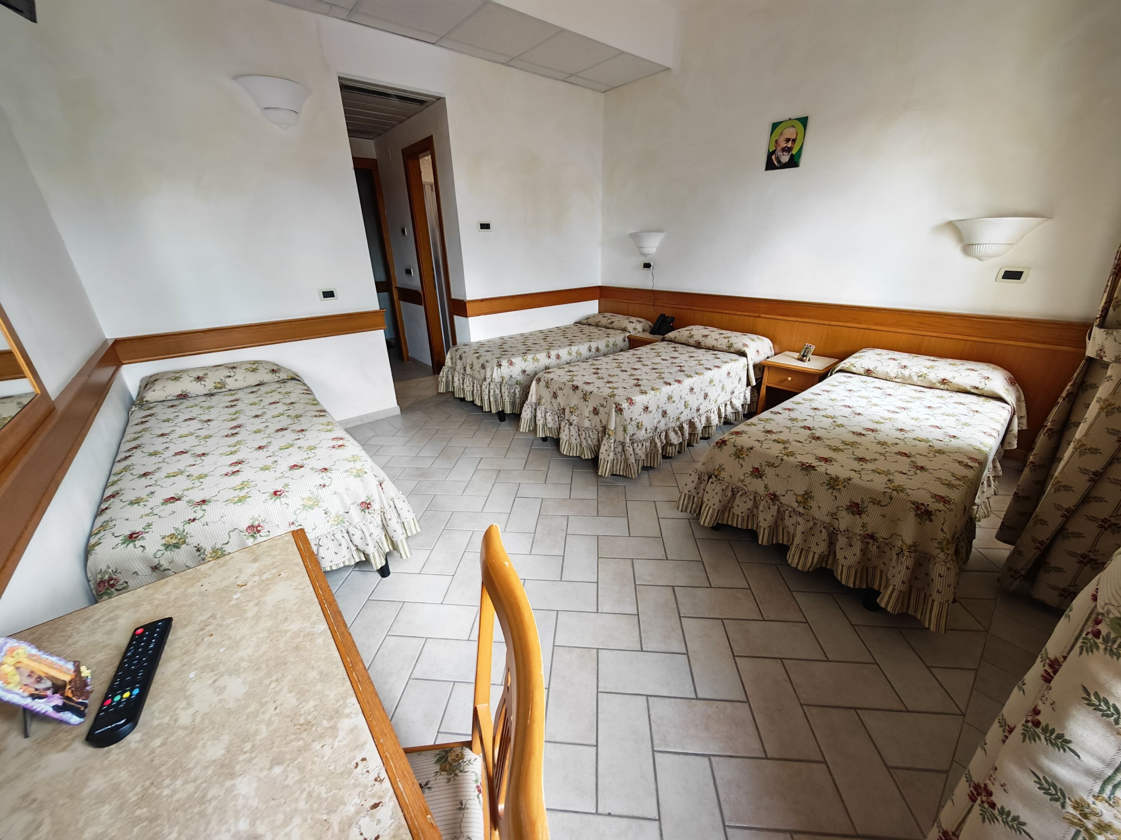 A hotel room with four single beds, a wooden headboard, a desk with a chair, a remote control, and a small table.