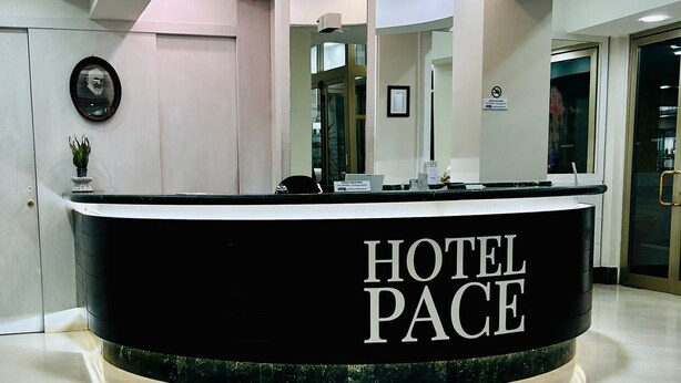 A hotel reception desk with a sign reading 'HOTEL PACE,' a framed picture on the wall, and a potted plant. - Hotel Pace (San Giovanni Rotondo)