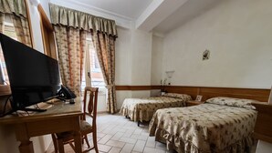A hotel room with a wooden desk, two beds with floral bedding, a television, and a window with curtains. - Hotel Pace (San Giovanni Rotondo)