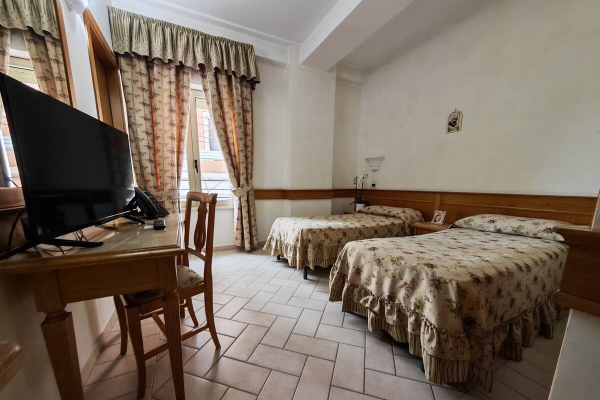A hotel room with a wooden desk, two beds with floral bedding, a television, and a window with curtains.