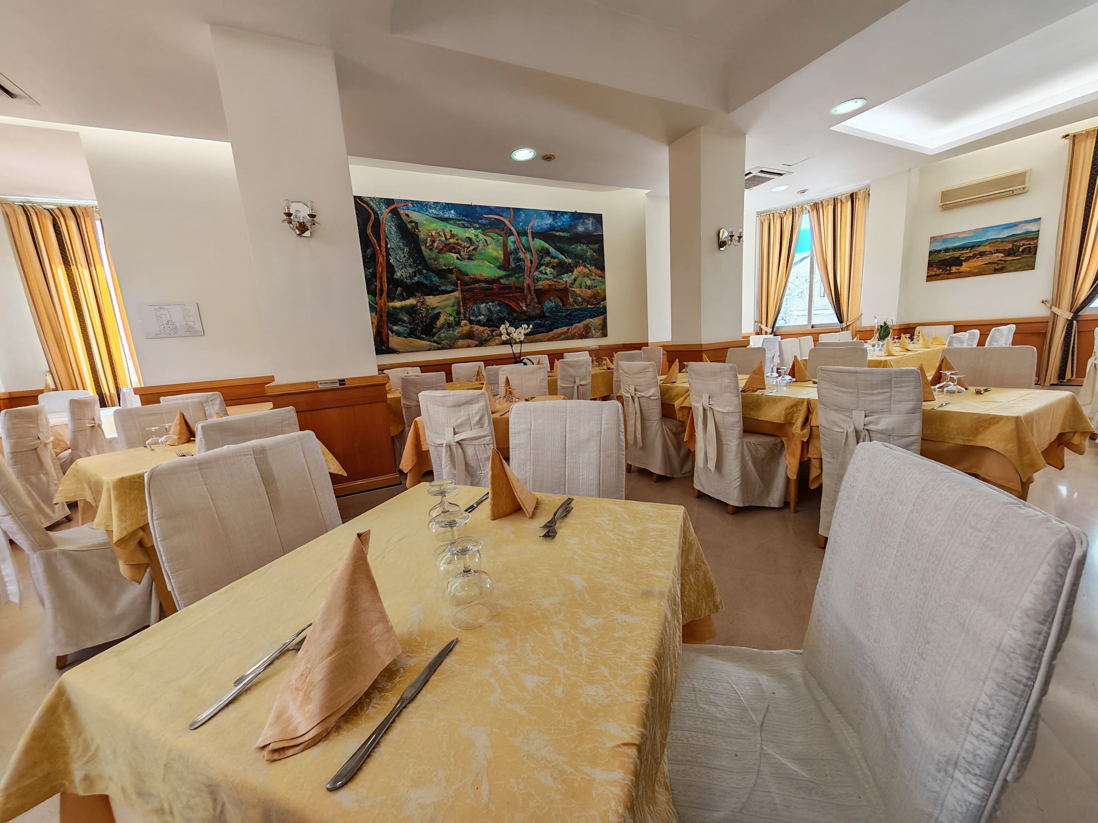 A dining area with tables set for a meal, chairs with white covers, and artwork on the walls.