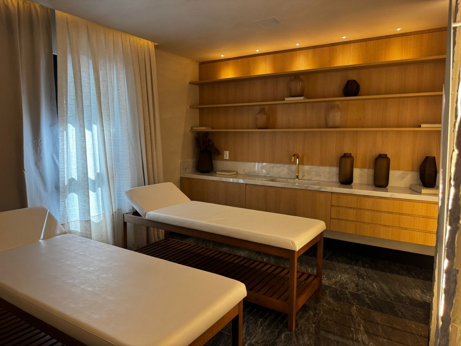 Um spa moderno com mesa de massagem, prateleiras de madeira com objetos decorativos e piso de mármore.