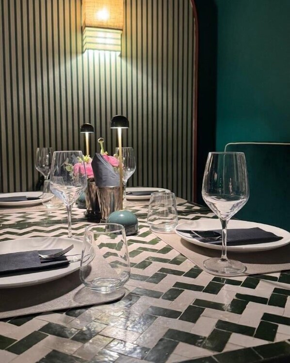A dining table set with plates, cutlery, and wine glasses, with a patterned tile floor and a green wall in the background.