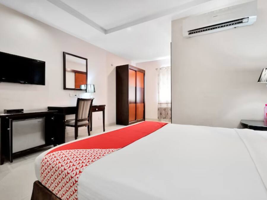 A hotel room with a bed, a desk, a chair, a TV, a wardrobe, and an air conditioning unit.