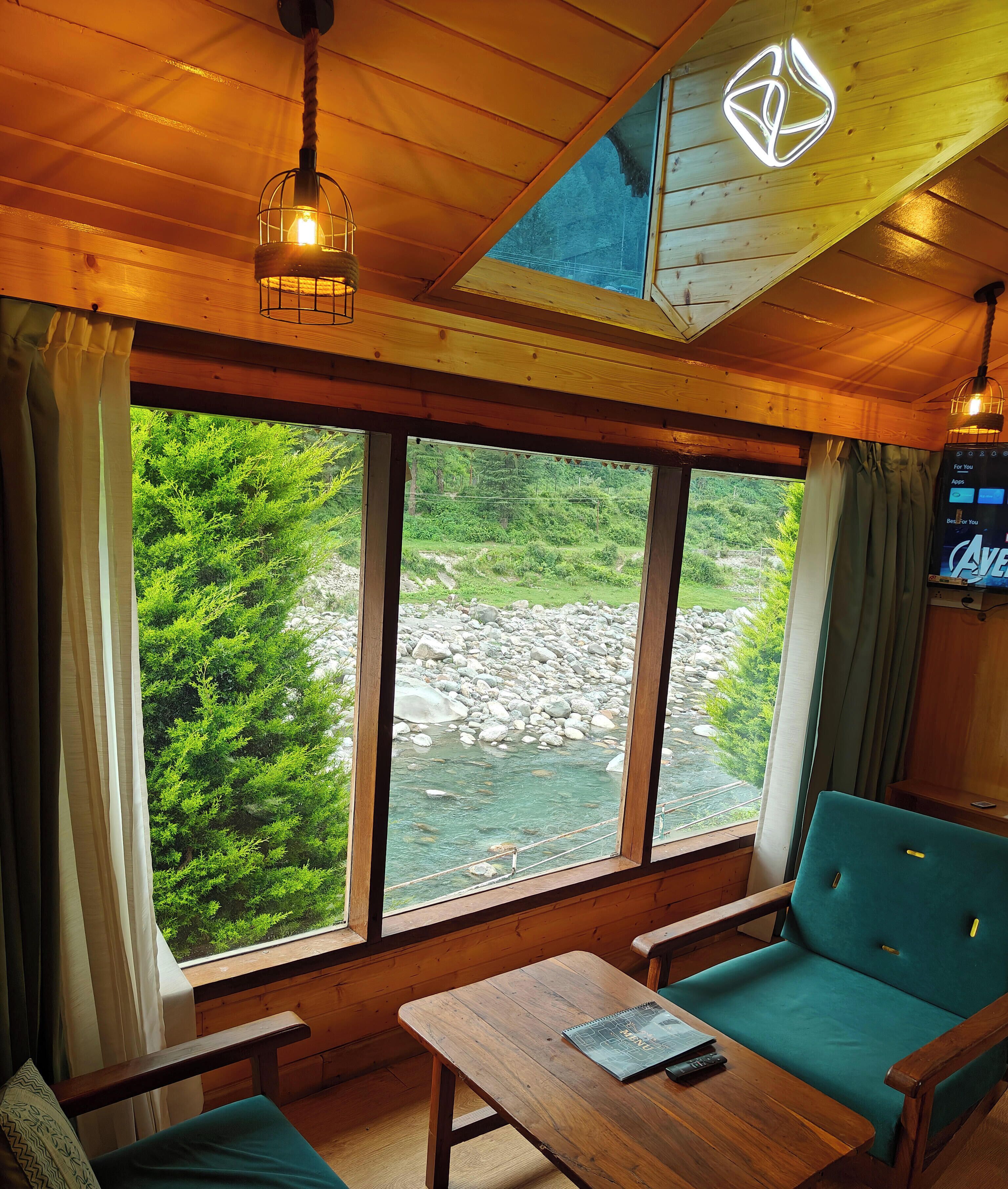 A cabin with a large window overlooking a river, a wooden ceiling, and a green sofa.