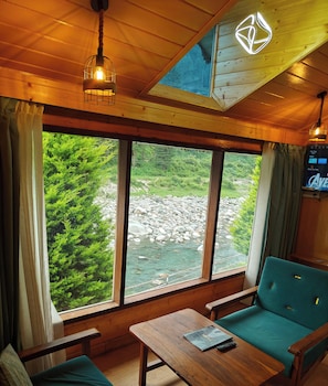 A cabin with a large window overlooking a river, a wooden ceiling, and a green sofa. - THE HEIMAT RESORT (Padhar)