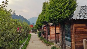 Un sendero de tierra estrecho bordeado por edificios de madera y vegetación exuberante, que conduce hacia un paisaje montañoso.