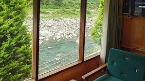 Una habitación con techo de madera, ventanales con vista a un río y follaje, una silla color turquesa y un televisor en la pared.